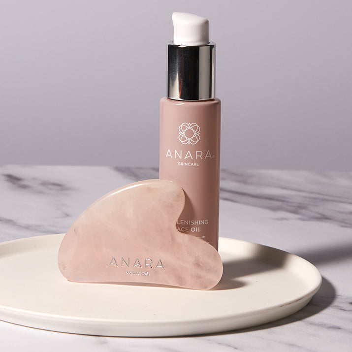 Anara Replenishing Face Oil with Rose Quartz Heart Shaped Gua Sha on a white plate and sitting on a marble surface.