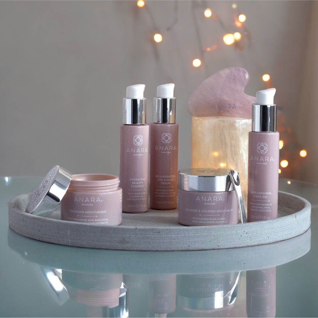 Radiance Moisturiser with the lid open, Cleanse & Nourish Night Balm, Rejuvenating Eye & Face Serum, Replenishing Face Oil, Hydrating Beauty Essence, Rose quartz Gua Sha placed on a stand. All placed on a grey tray with dots of light in the background.
