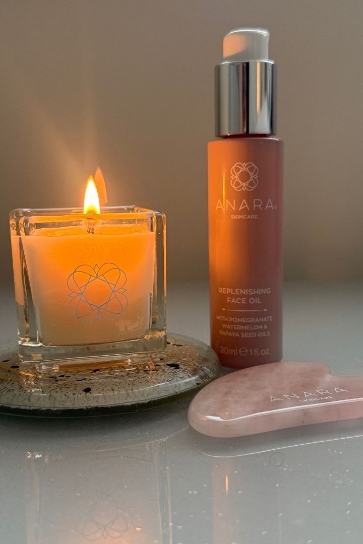 Anara skincare bottle, candle and Gua Sha on a reflective surface with a warm glow.